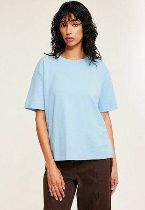 Vrouw met schouderlang golvend haar, gekleed in een lichtblauw kort mouw shirt en donkerbruine broek, staande voor een effen achtergrond.