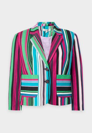 Høj person iført farverig, vertikalt striped blazer og matchende brede bukser med sort top og sorte sko, stående mod hvid baggrund.