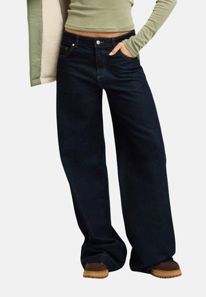 Person trägt hochgeschnittene, weit geschnittene, dunkelblaue Jeans mit der Hand in der Tasche, olivgrünes Langarmoberteil und braune Schuhe vor weißem Hintergrund.