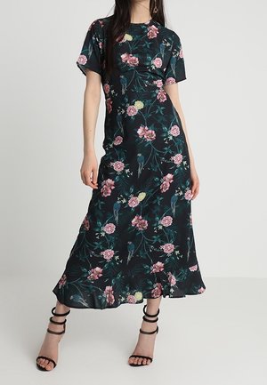 Frau trägt ein schwarzes Midikleid mit Blumenmuster, kurzen Ärmeln und schwarze High-Heels mit Riemchen vor schlichter Hintergrund.