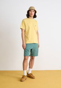T-shirt en coton jaune avec une poche sur la poitrine, associé à un short teal et des chaussures à lacets marron. L'arrière-plan est de couleur claire.