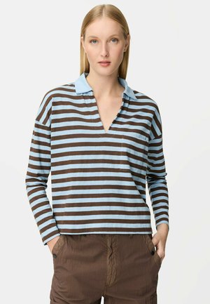 Frau trägt ein blau-braun gestreiftes Langarmshirt mit blauem Kragen und brauner Hose, steht mit den Händen in den Taschen.