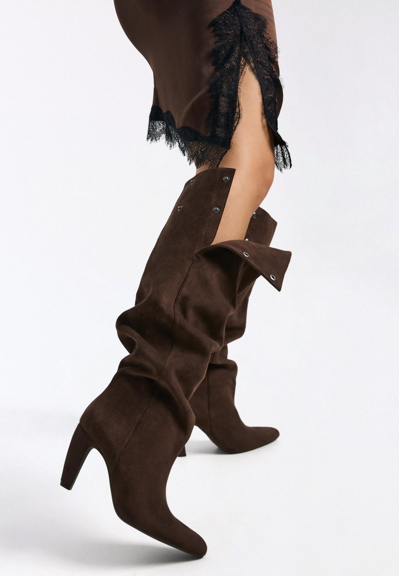 Bottes marron en daim jusqu'aux genoux avec un bout pointu, un design ample, une fermeture à pression et un talon haut et fin.