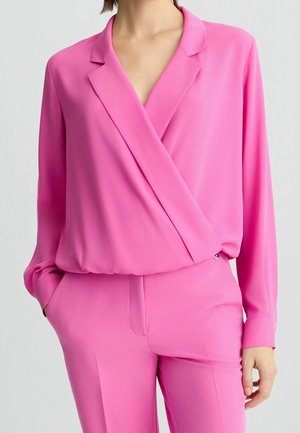 Frau trägt eine leuchtend pinke Wickelbluse mit langen Ärmeln und passende hoch taillierte Hose, eine Hand in der Tasche, vor schlichtem Hintergrund.