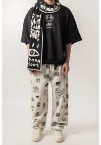 Czarna, oversize koszulka z białą grafiką, wzorzysty szalik, kremowe spodnie z czarnymi rysunkami i białe sneakersy. Materiał wydaje się miękki i wygodny.