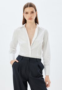 Camisa de cuellos blancos con botones al frente, que presenta un sutil bordado en el pecho, combinada con pantalones marino a rayas y detalle en los bolsillos.