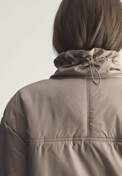 Person trägt taupefarbene gesteppte Jacke mit hohem Kragen und verstellbarem Kordelzug, von hinten vor einfachem Hintergrund gesehen.
