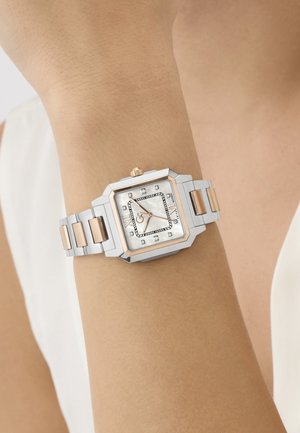 Vierkante zilveren en roségouden polshorloge met diamanten uurmarkeringen op de pols van een persoon tegen een neutrale achtergrond.