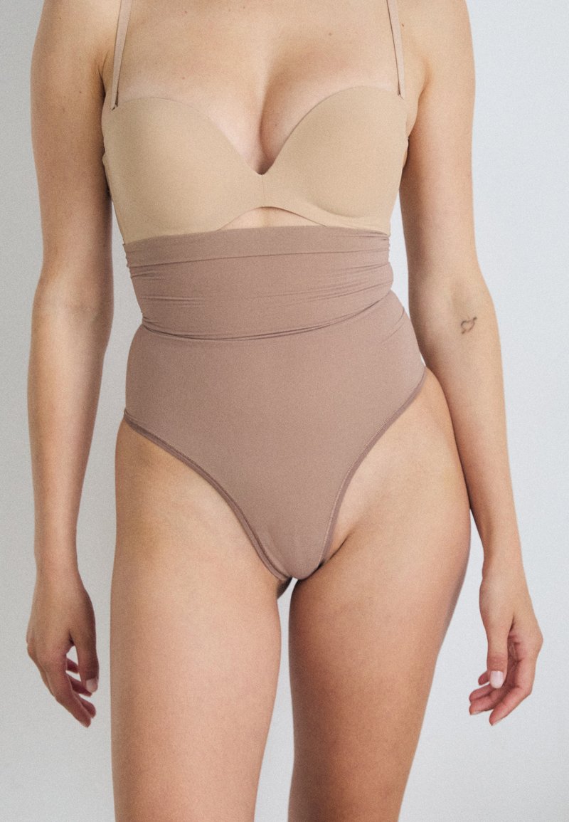 Bodysuit featuring a strapless nude bra top, high-waisted beige fabric with ruching, smooth texture, and high-cut legs.