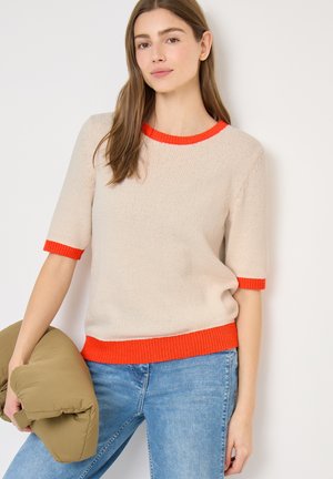 Młoda kobieta w beżowym swetrze z jaskrawym pomarańczowym obszyciem, niebieskich dżinsach, trzymająca oliwkowozieloną kurtkę puchową na białym tle.