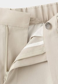 Ceinture de pantalon beige montrant une fermeture éclair, un bouton et une bande élastique intérieure ajustable pour un ajustement personnalisé.