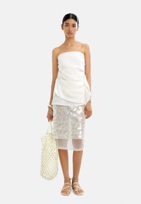 Biała top bez ramiączek z drapowanym materiałem, zestawiona z przezroczystą spódnicą do kolan pokrytą cekinami. Modelka trzyma plecioną beżową torbę i ma na sobie sandały na obcasie.