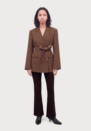 Femme debout portant un blazer marron ceinturé, un pantalon évasé marron foncé à motif léopard et des bottines noires à talons sur un fond uni.