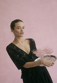 Schwarzes Spitzentop mit kurzen Ärmeln, hält ein silbernes Tablett mit fünf rosa Getränken in Kristall-Coupette-Gläsern vor einem sanften rosa Hintergrund.