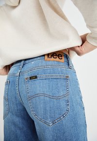 Sinine denimist teksad, millel on beež nahkabelt, millel on "Lee" logo, esiletõstetud õmblused ja eristuva tasku detailid.