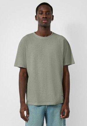 Young man with dark skin wearing a plain olive green short-sleeve t-shirt and light blue jeans, standing against a white background.