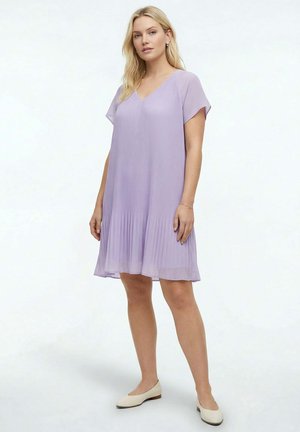 Femme debout portant une robe plissée à manches courtes, de couleur violet clair, et des chaussures plates beiges, sur un fond blanc uni.