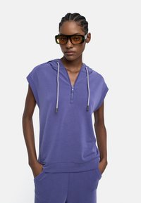 Osoba nosząca fioletowy bezrękawnik z zamkiem i sznurkiem, pasujące spodnie oraz duże prostokątne brązowe okulary przeciwsłoneczne.