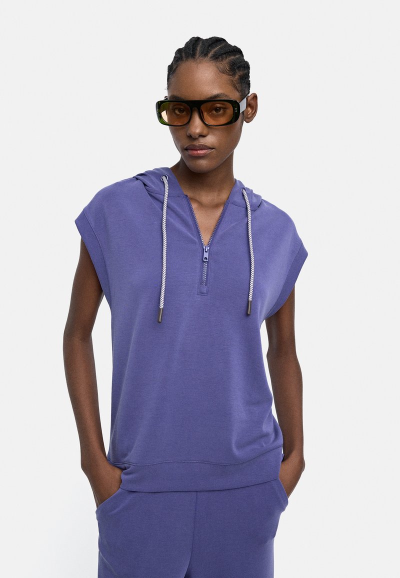 Osoba nosząca fioletowy bezrękawnik z zamkiem i sznurkiem, pasujące spodnie oraz duże prostokątne brązowe okulary przeciwsłoneczne.