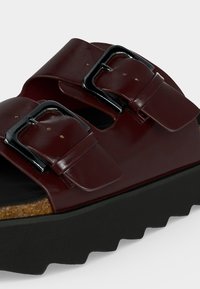 Σανδάλι από σκούρο κόκκινο δέρμα με δύο ρυθμιζόμενα ασημένια αγκράφες και παχύ μαύρο πάτο πλατφόρμας με κυματιστό πέλμα.