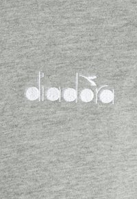 Logo "diadora" brodé en blanc avec un petit détail de feuille au-dessus du "o" sur un fond de tissu gris clair.