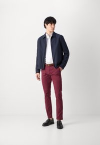 Marineblaues Bomberjackett, weißes Hemd, bordeauxfarbene Hose und schwarze Slipper. Glatte Textur, Reißverschluss und ein brauer Gürtel, der den Look betont.
