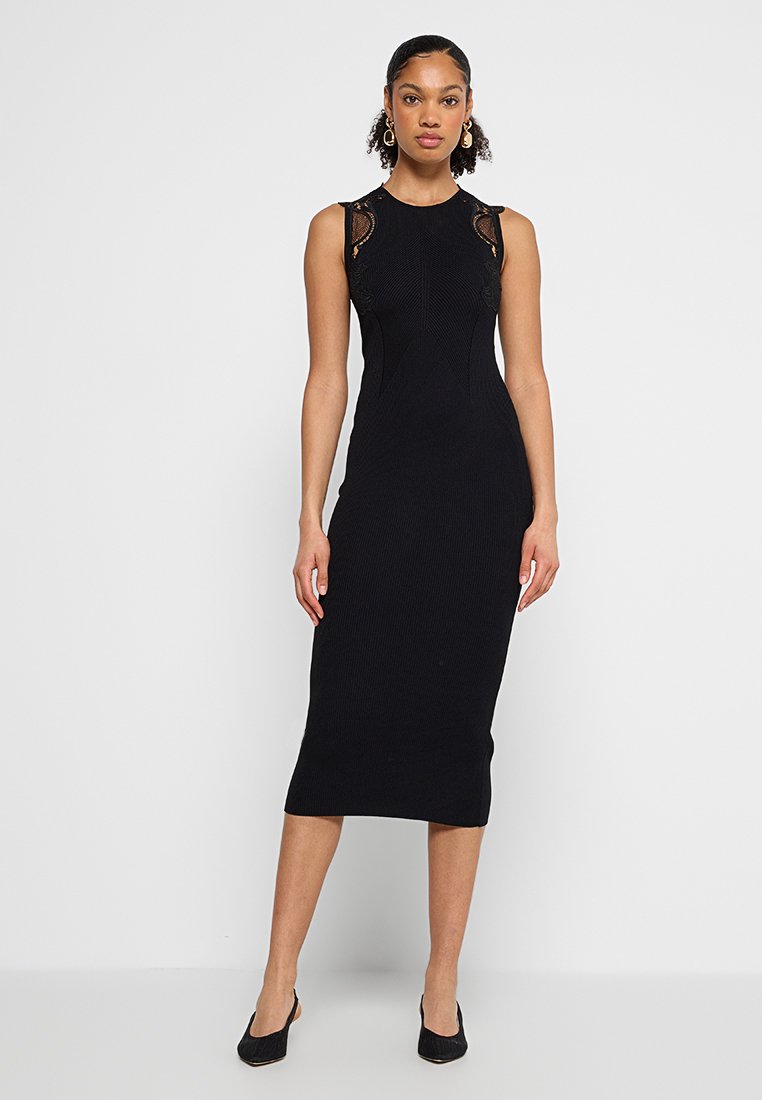 Czarna bezrękawnikowa sukienka z prążkowanego materiału, z dopasowanym gorsetem, gładką fakturą, długości mid-calf, z misternymi koronkowymi akcentami na ramionach, zestawiona z czarnymi butami.
