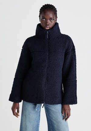 Giovane con capelli intrecciati, che indossa una giacca zip-up blu navy a trama e jeans azzurri chiari, in piedi contro uno sfondo semplice.