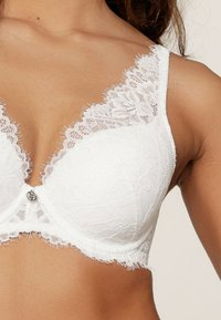 Soutien-gorge blanc en dentelle avec des bonnets rembourrés et un motif floral. Dispose d'une bordure en dentelle sur les bretelles et d'un fermoir en métal décoratif au centre.