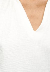 Gros plan d'une personne portant un haut blanc texturé à col en V, montrant une partie du cou et du haut de la poitrine.