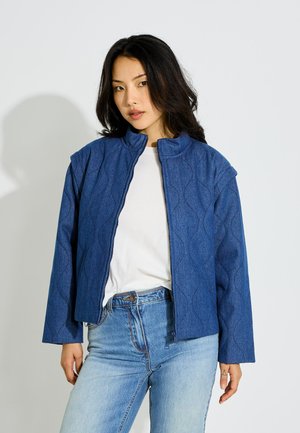 Joven mujer que lleva una chaqueta acolchada azul sobre una camisa blanca y vaqueros azules claros, de pie frente a un fondo claro y liso.