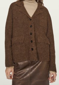 Donna che indossa un cardigan marrone morbido con bottoni sopra un dolcevita beige, abbinato a una gonna in pelle marrone con texture a coccodrillo e anelli in argento.