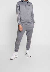 Svetle šedý sportovní komplet Nike zahrnující zipovou mikinu s šňůrkou, odpovídající jogger kalhoty a kontrastní černé pruhy po stranách. Bílé tenisky.