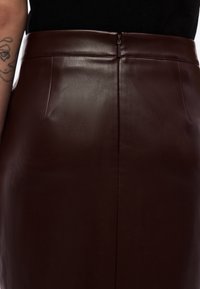 Καφέ δερμάτινη φούστα με ομαλή υφή, ψηλή μέση, κλείσιμο με φερμουάρ στο πίσω μέρος και δύο διακριτικές πλαϊνές ραφές. Μινιμαλιστικός σχεδιασμός, χωρίς ορατά σχέδια.