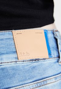 Svijetloplave traperice s tamnom kožnom etiketom s utisnutim tekstom. Šavovi su u kontrastnoj narančastoj boji.