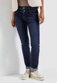 Jeans en denim bleu foncé avec une taille mi-haute, fermeture à deux boutons, design à cinq poches et ourlets retroussés, assortis à des baskets beige et rouge.