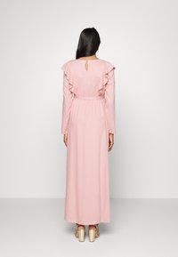 Vestido longo rosa com mangas com babados, textura trançada e detalhe de abertura nas costas. O tecido transparente desce até o tornozelo, combinado com sapatos de salto dourados.