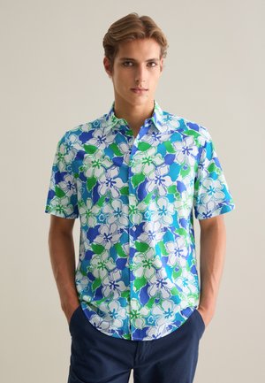 Camisa de manga corta en azul con un patrón floral en verde y blanco. Hecha de tela ligera, presenta un collar abotonado y un dobladillo redondeado.