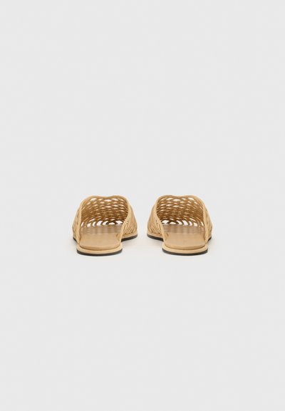 Sandales ouvertes à bout en cuir clair, avec un design tressé et une semelle plate. Le matériau semble flexible avec des motifs découpés pour une meilleure aération.