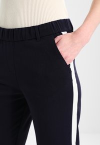 Granatowe spodnie o gładkiej fakturze, z białym paskiem po boku i elastycznym paskiem w talii. Zauważalna kieszeń boczna na wysokości biodra.