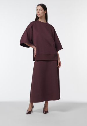 Femme portant un haut ample de couleur bordeaux foncé avec des manches jusqu'au coude et une longue jupe assortie, associée à des chaussures à talons hauts foncées, debout.