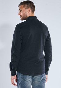 Schwarzes Langarmhemd mit einer weichen Textur, flachem Kragen und Knopfmanschetten, von hinten gezeigt, kombiniert mit hellblauen Jeans.