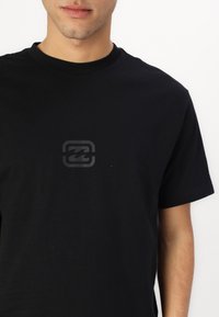 Černé bavlněné tričko s kulatým výstřihem a krátkým rukávem, uprostřed s jemným šedým logem. Měkká textura a jednoduchý design.