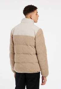 Veste matelassée avec un corps en polaire beige et un haut en nylon beige clair. Caractéristiques : col haut et surface texturée avec des sections matelassées.