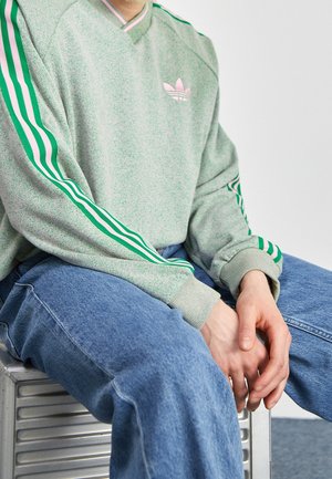 Osoba siedząca na metalowej ławce ma na sobie zieloną bluzę Adidas z pasiastymi rękawami i niebieskie jeansy, ręce spoczywają na kolanach.
