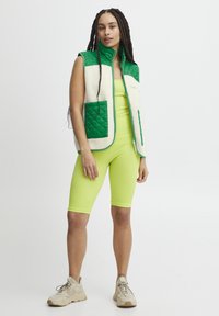 Chaleco bloqueado en verde y crema con diseño acolchado, cuerpo de forro polar texturizado y bolsillos laterales, llevado sobre un conjunto de canalé verde brillante.