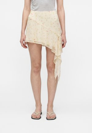 Kvinde iført en cremet blomsterasymmetrisk mininederdel med flæsedetalje kombineret med beige tanga-sandaler.