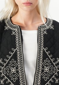 Black quilted jacket featuring intricate white embroidery along the neckline and front, with a soft texture and fitted design.
