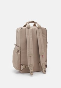 Sac à dos beige avec un dos et des bretelles rembourrés, en forme rectangulaire, poches latérales et matériel ajustable. Fabriqué en tissu durable.