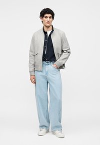 Νέος άνδρας με σγουρά μαλλιά που φορά γκρι bomber μπουφάν, μαύρο πουκάμισο, ανοιχτό μπλε τζιν με φαρδιά πόδια και λευκά αθλητικά παπούτσια, στέκεται με το ένα χέρι στην τσέπη.